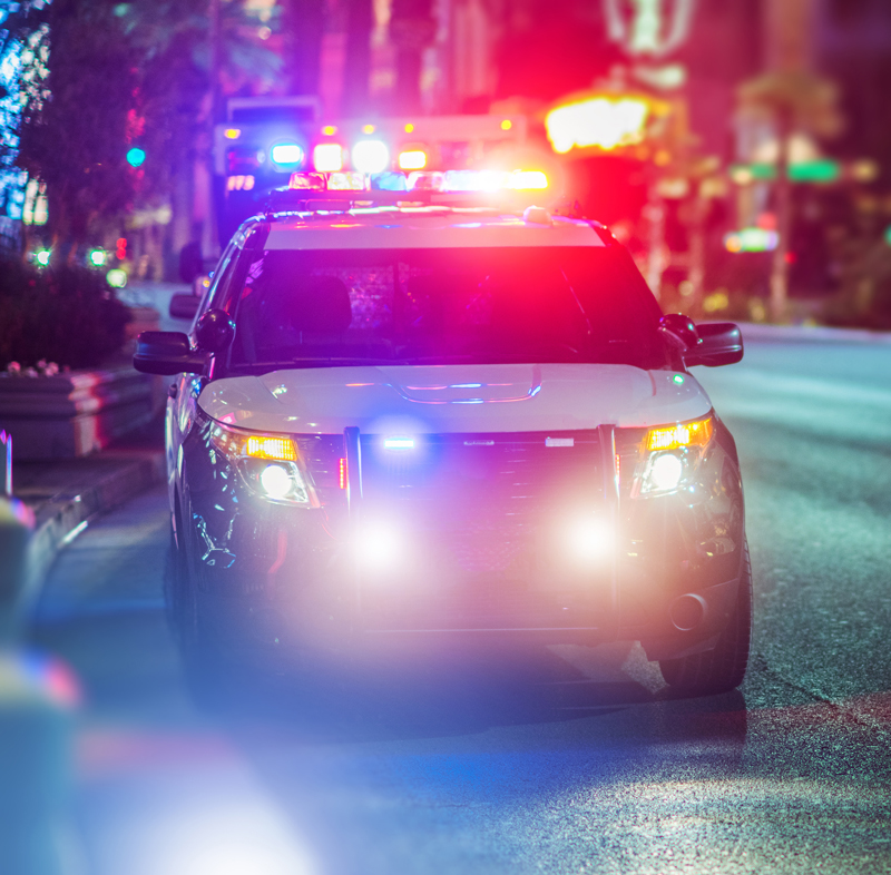 Police car with flashing lights parked on a city street at night, with blurred lights and an emergency vehicle visible in the background.