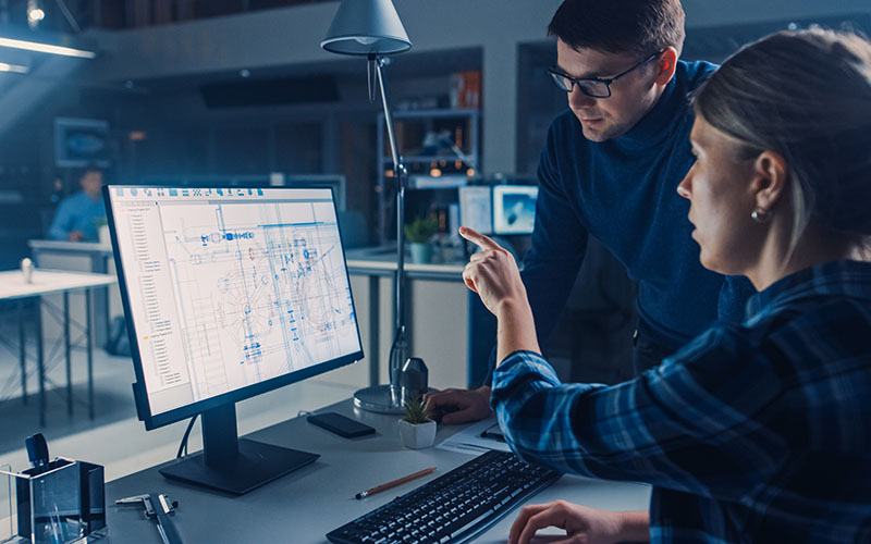 Deux personnes dans un bureau regardent un écran d'ordinateur affichant des schémas techniques. L'une des personnes pointe l'écran tandis que l'autre se tient à côté d'elle.