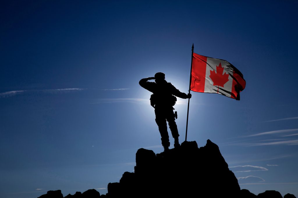 Un soldat se tient sur un pic rocheux, saluant à côté d'un drapeau canadien dans un ciel bleu et clair, avec le soleil derrière eux.