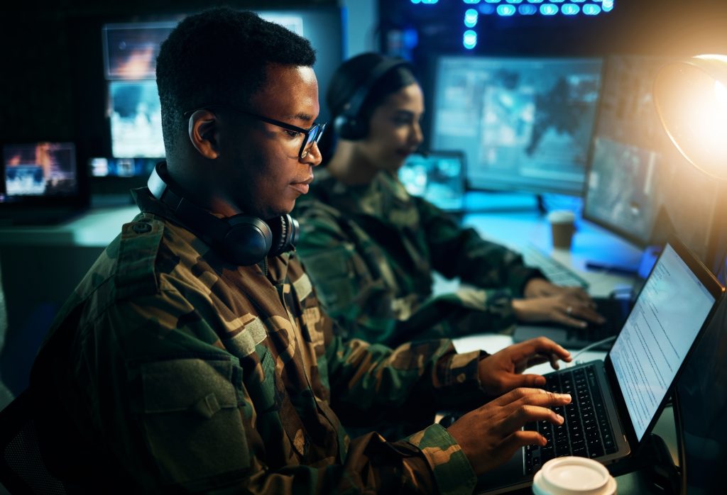 Deux personnes en uniforme militaire travaillent sur des ordinateurs portables dans une pièce faiblement éclairée, avec plusieurs écrans d'ordinateur et des équipements en arrière-plan.