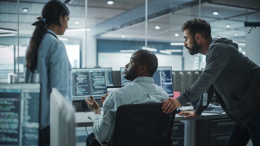 Trois personnes discutent d'un code affiché sur des écrans d'ordinateur dans un bureau moderne, une personne assise et deux personnes debout à proximité, toutes concentrées sur les écrans.