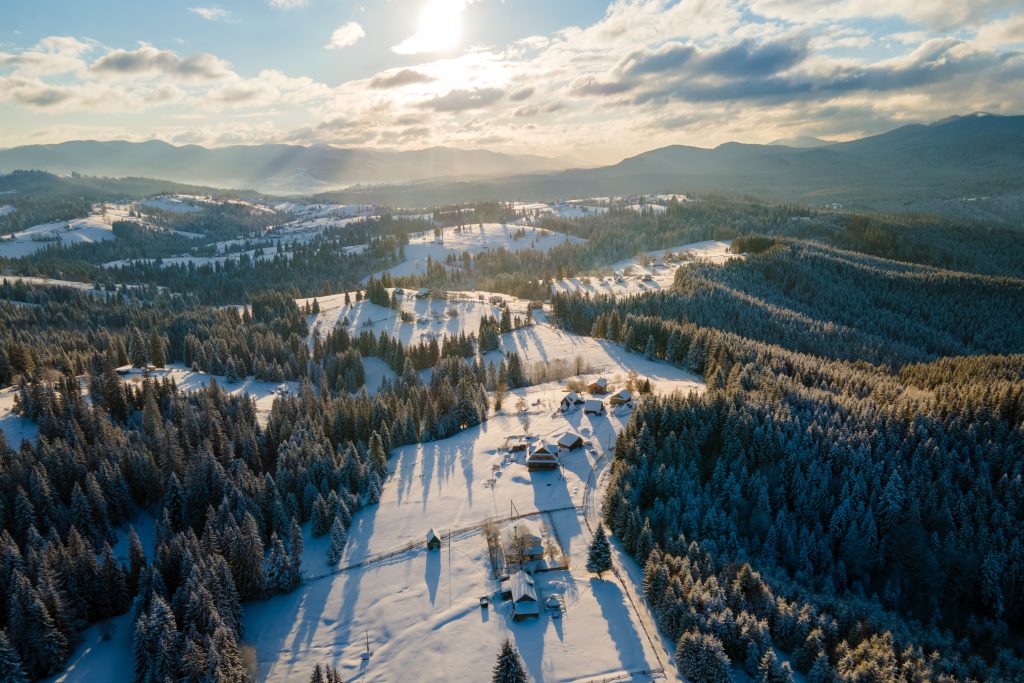 Vue aérienne d'un paysage rural enneigé avec des maisons dispersées, des forêts denses à feuilles persistantes et des montagnes lointaines sous un ciel partiellement nuageux au lever ou au coucher du soleil.