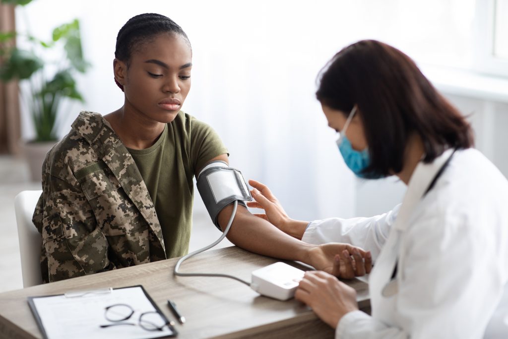 A healthcare professional measures the blood pressure of a woman in military uniform using a digital monitor at a medical office.