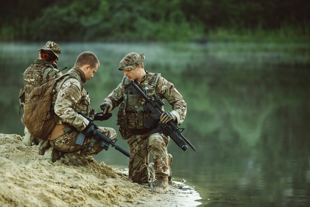 Trois soldats en tenue de camouflage : deux d'entre eux sont à genoux sur une plage de sable, un fusil à la main, et le troisième est à genoux près de l'eau ; on aperçoit des arbres et de la végétation en arrière-plan.