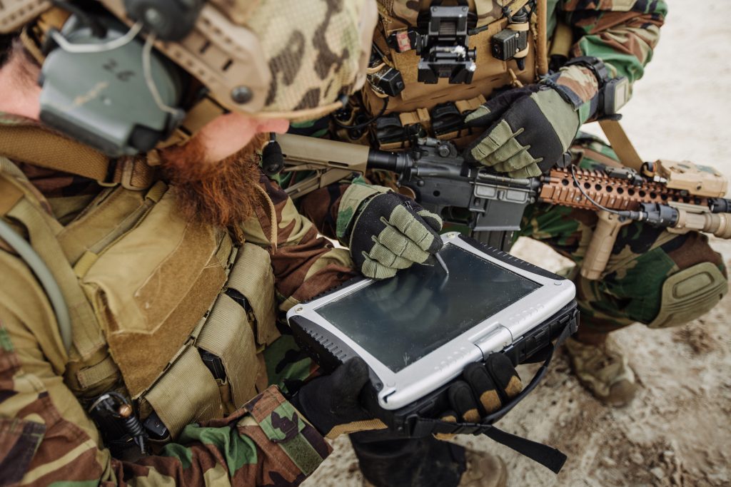 Deux soldats en tenue de camouflage et équipement tactique utilisent une tablette robuste à l'extérieur ; l'un d'eux tient un fusil.
