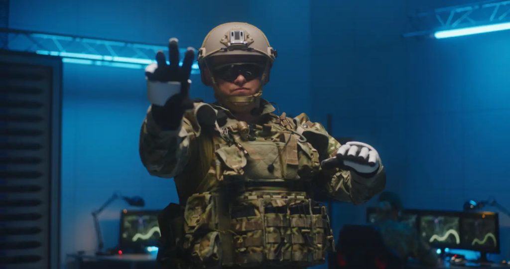 Un soldat en tenue de combat et portant un casque se tient à l'intérieur, une main levée, avec des écrans d'ordinateur et du matériel visibles à l'arrière-plan.