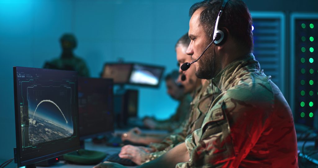 Dans une salle de contrôle, des militaires équipés de casques d'écoute surveillent des écrans d'ordinateur affichant les trajectoires de vols ou de missiles.