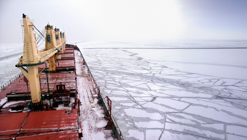 Un grand cargo navigue dans des eaux glacées, brisant des plaques de glace sous un ciel nuageux.