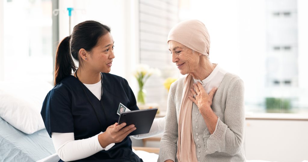 Un professionnel de la santé montre une tablette à une femme âgée portant un foulard, dans une chambre d'hôpital.