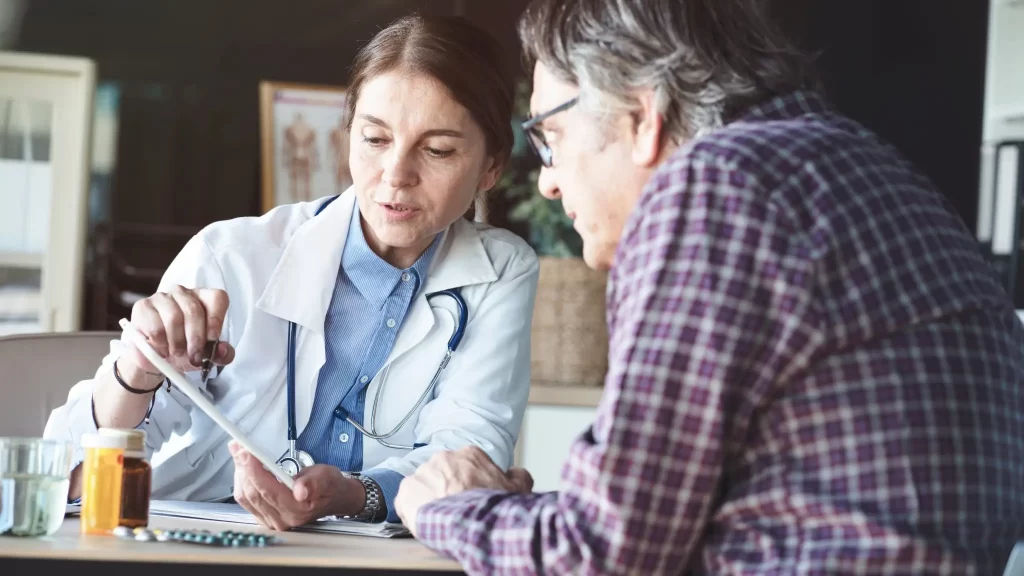 A doctor shows information on a tablet to a patient sitting across the table, with medicine bottles and a glass of water nearby.