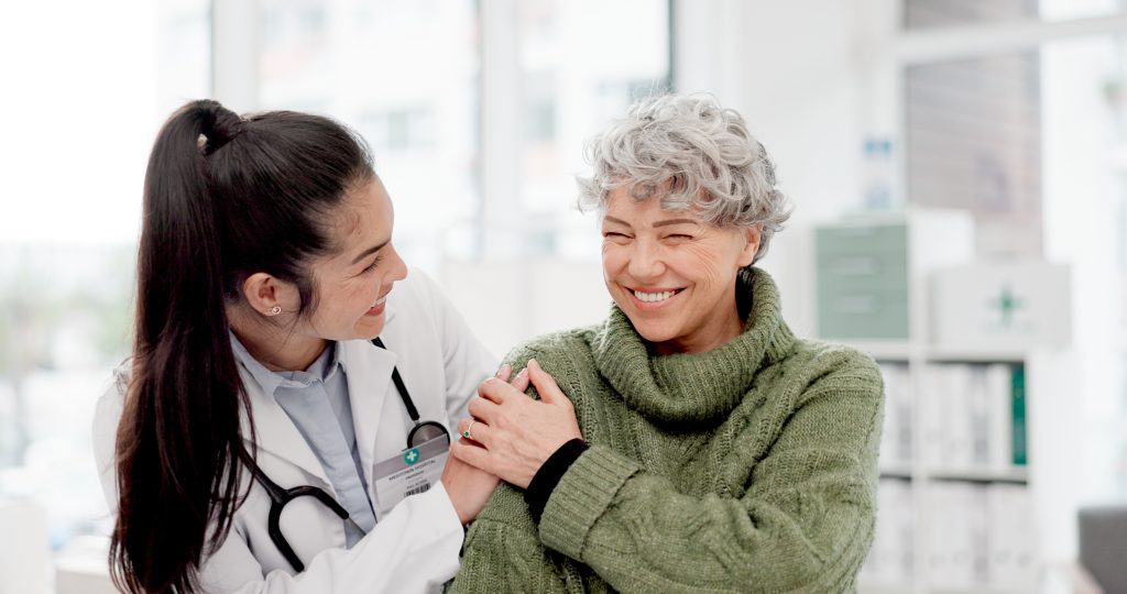 Un médecin en blouse blanche sourit et tient doucement l'épaule d'une femme plus âgée en pull vert, qui sourit également, dans un cabinet médical.