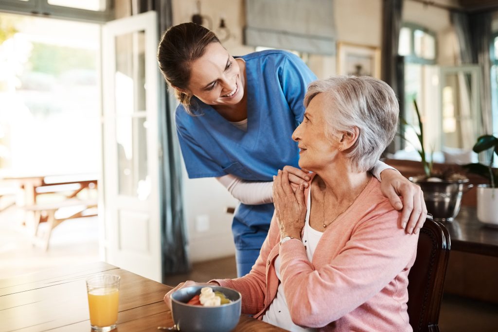 A caregiver in blue scrubs smiles and talks with an older woman sitting at a table with a bowl of food and a glass of orange juice.
