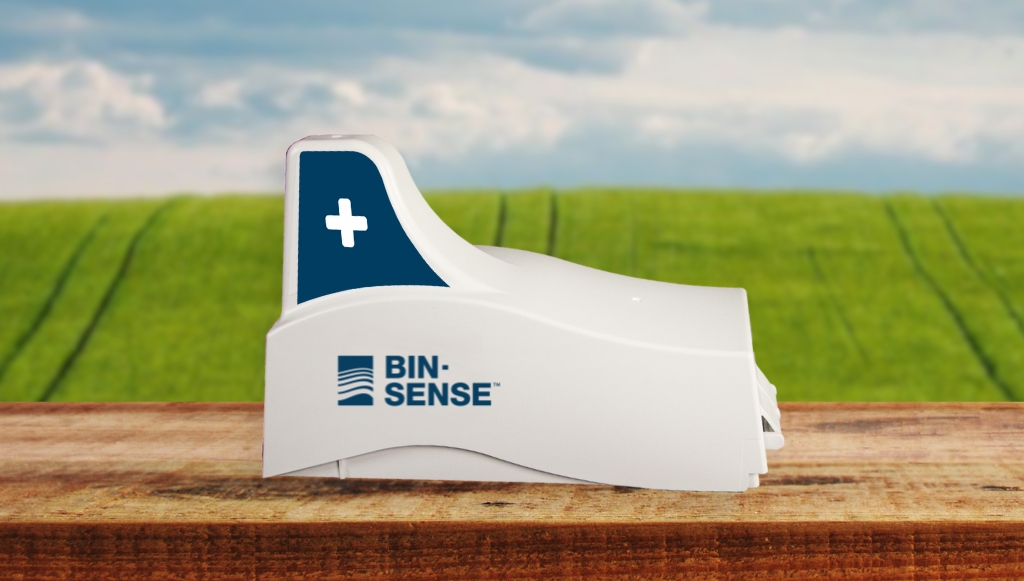A white device labeled "BIN-SENSE" with a blue cross symbol, placed on a wooden surface, with a background of a grassy field and cloudy sky.