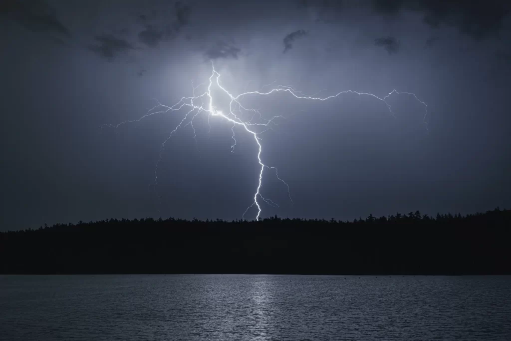 Lightning striking over a lake at night with dark silhouetted trees along the shoreline and a cloudy sky.