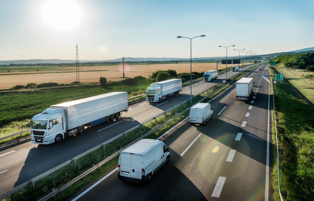 Aerial view of a highway with multiple trucks and vans driving in both directions on a clear day. Fields and hills are visible in the background.