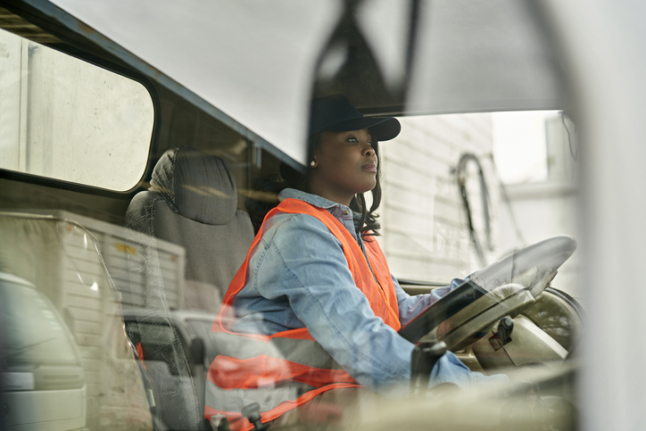 A person wearing a black hat, orange safety vest, and blue shirt is driving a vehicle, as viewed through the windshield from outside.