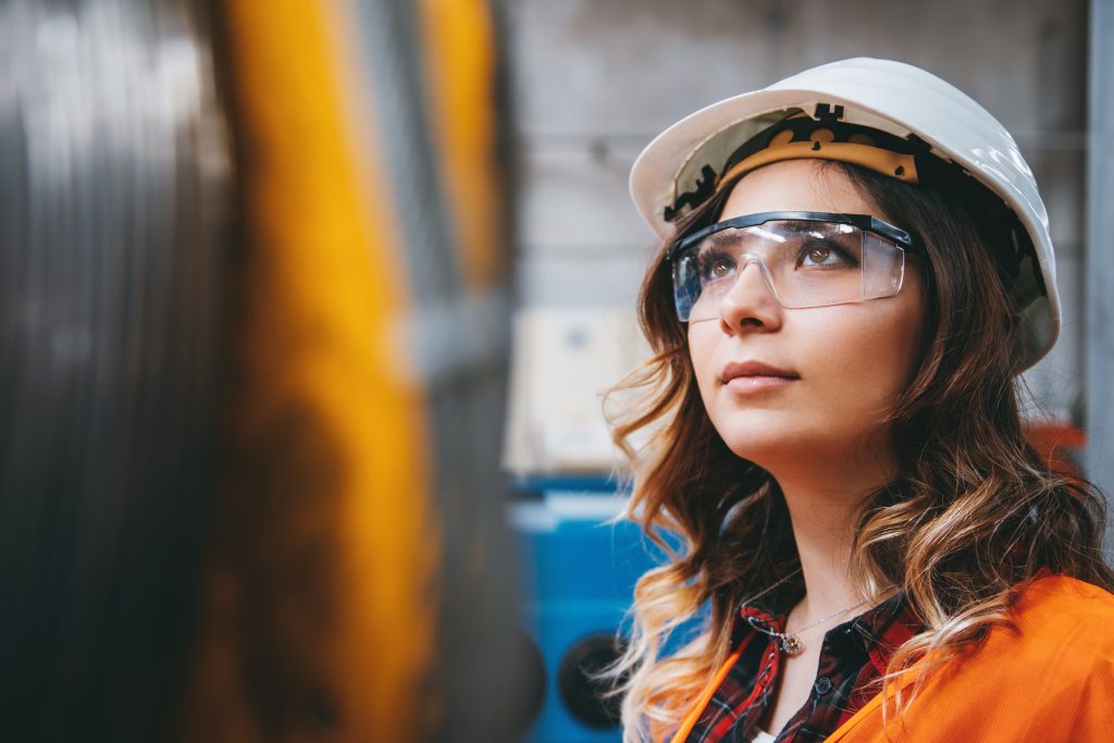 Une femme portant un casque et des lunettes de sécurité regarde en l'air, debout dans un environnement industriel avec des machines floues au premier plan.