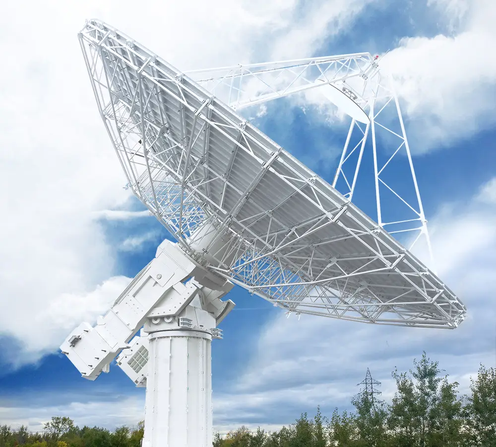 A large white satellite dish antenna is positioned outdoors against a partly cloudy blue sky, surrounded by green trees and power lines in the background.