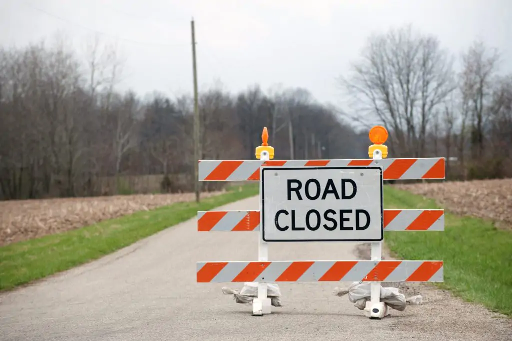 A "Road Closed" sign blocks a rural road, with orange and white barricades and warning lights. Trees without leaves and fields are visible in the background.