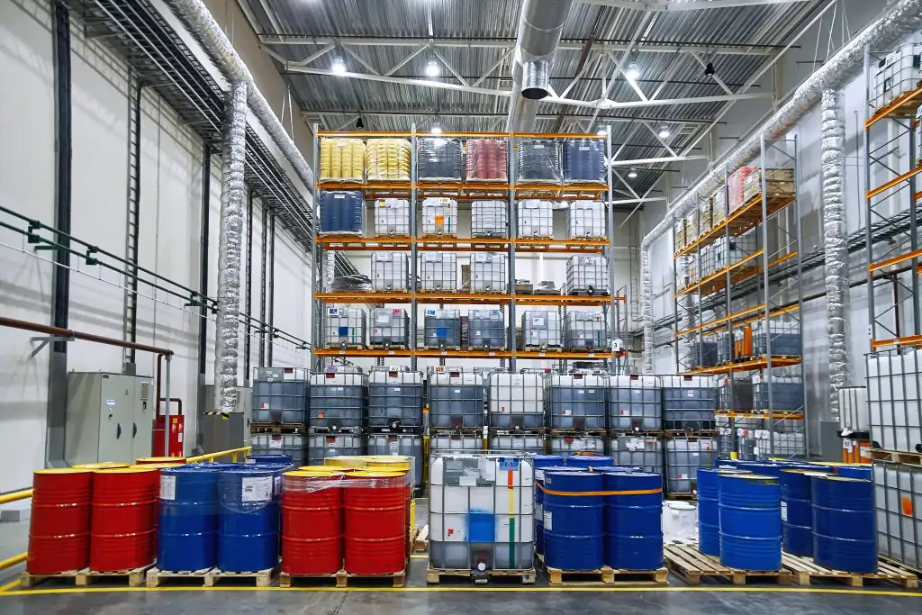 Industrial warehouse with shelves storing various large barrels and containers, including red, blue, and yellow drums, under bright overhead lighting.