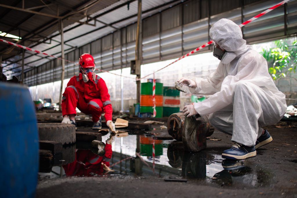 Two people in protective suits and masks clean up debris and contaminated items near a puddle inside an industrial building.