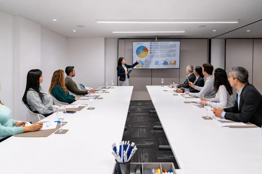 A woman presents data charts on a screen to a group of people seated around a long conference table in a modern meeting room.