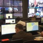 People work at computer stations in a control room with large screens on the wall displaying maps, data, and a video call.