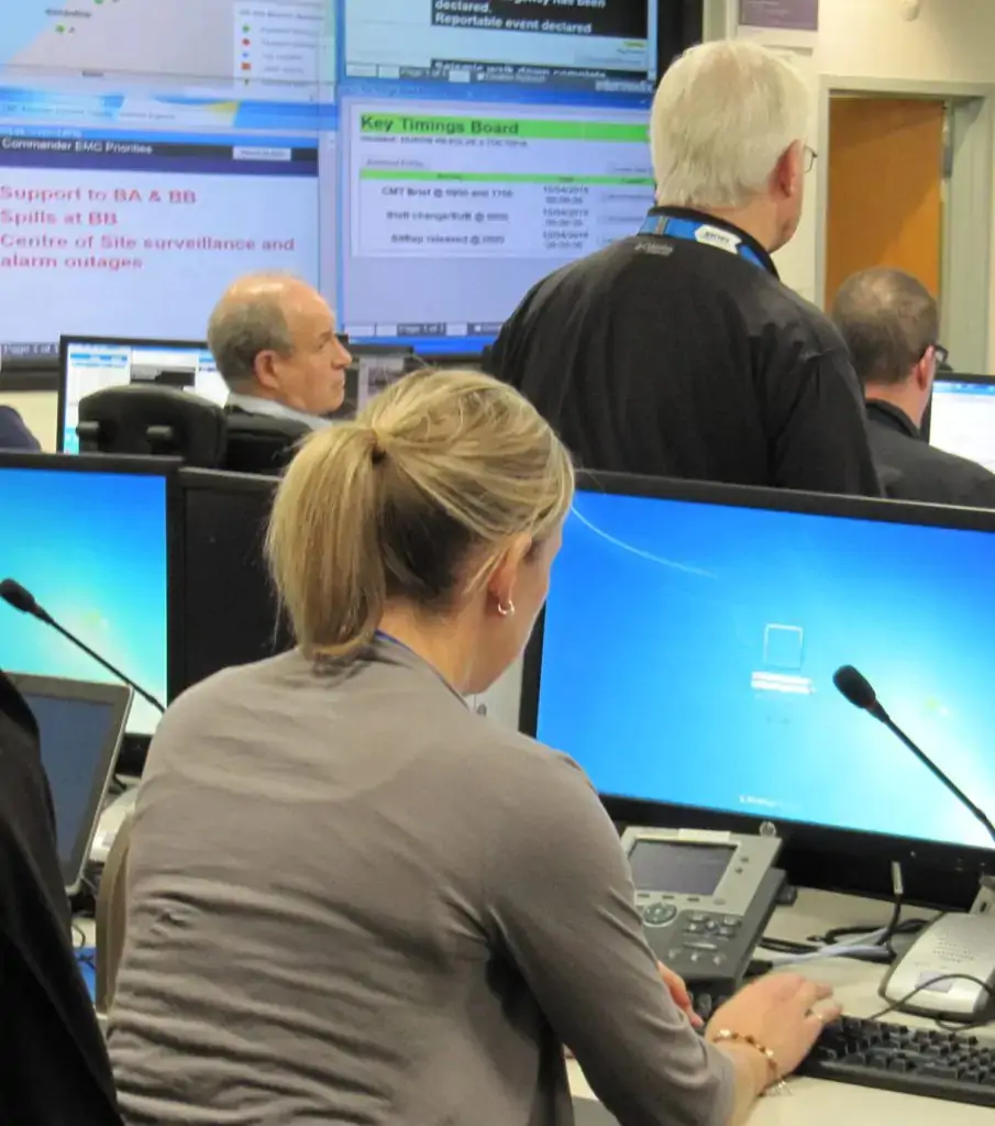 People working at computer stations in an office, with several large display screens showing data and schedules on the wall.