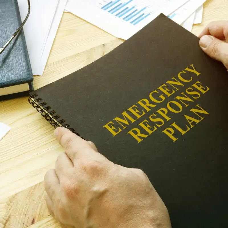 A person holds a black spiral-bound folder labeled "Emergency Response Plan" on a wooden desk with scattered papers and a blue notebook.