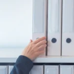 A person’s hand reaching for a white binder on a shelf filled with other similar binders.