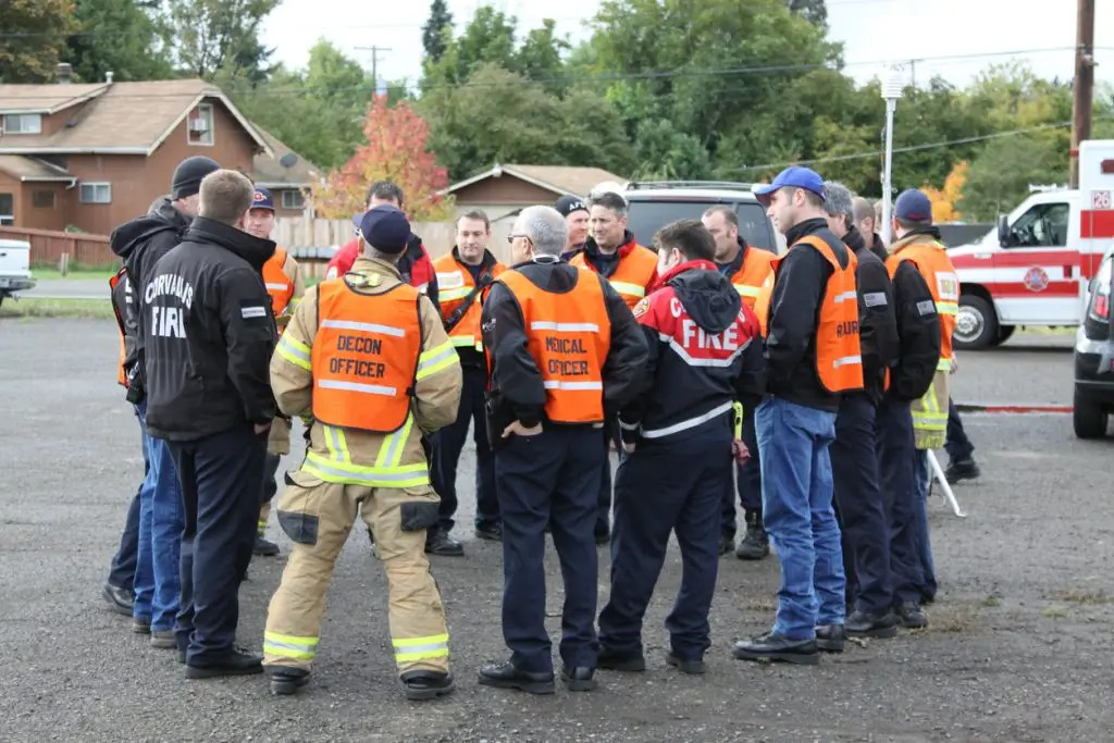 A group of emergency responders wearing safety vests and uniforms stand in a circle outdoors, appearing to discuss a plan near emergency vehicles.
