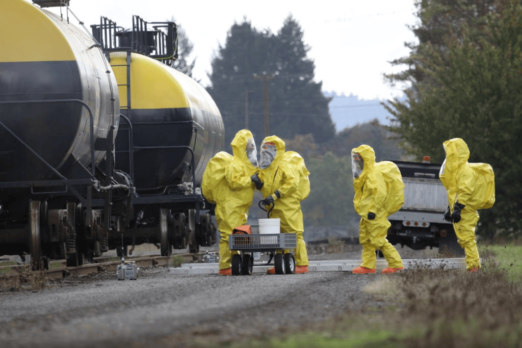 Four people in yellow hazmat suits work beside train tank cars on a gravel path, with equipment placed on a cart in front of them.