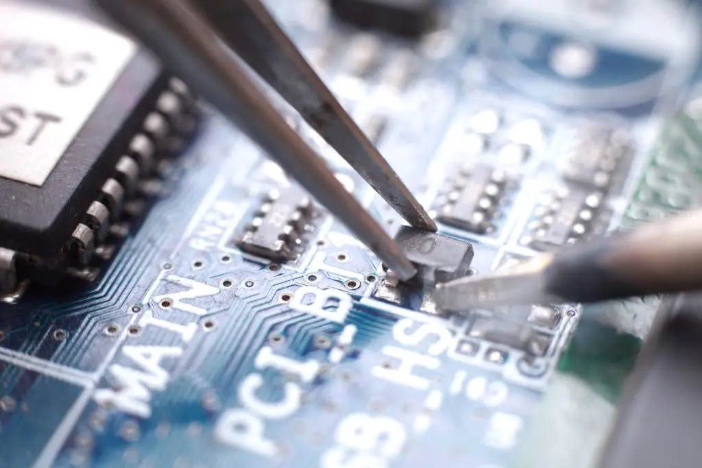 Close-up of tweezers placing a small electronic component onto a blue printed circuit board with visible microchips and labels.