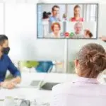 Three people sit at a conference table while participating in a video call with four others displayed on a screen; one in-person attendee wears a face mask.