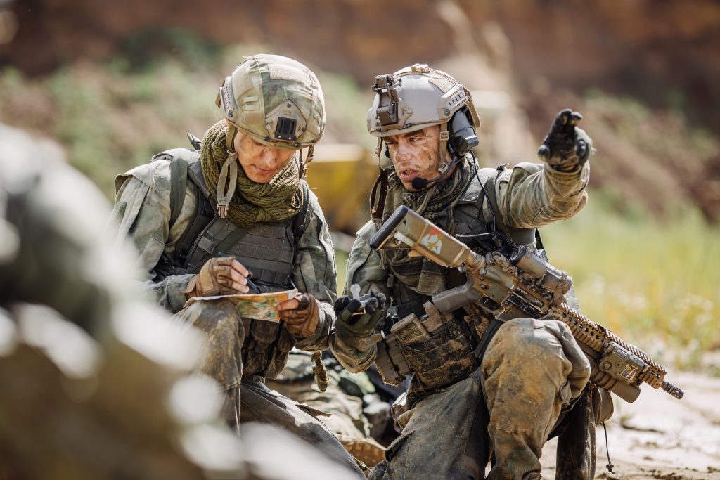 Two soldiers in camouflage gear study a map; one writes while the other points toward something off-frame, both wearing helmets and tactical equipment.