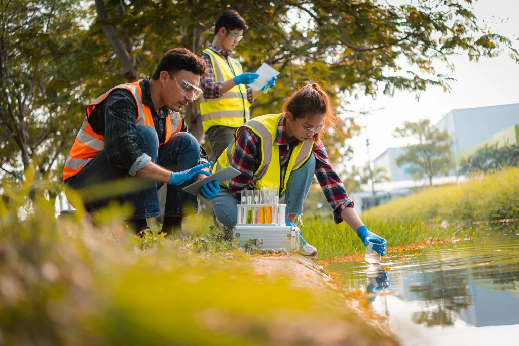 Trois personnes en tenue de sécurité prélèvent des échantillons d'eau au bord d'une rivière, à l'aide d'éprouvettes, et enregistrent les données en vue d'une analyse environnementale.