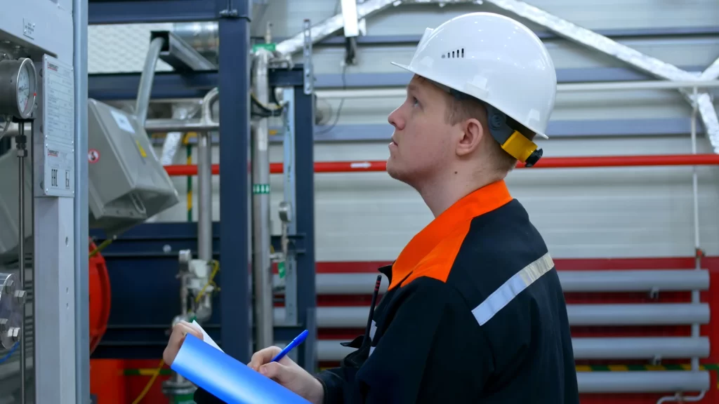 Un ouvrier portant un casque blanc et une veste de sécurité inspecte l'équipement et prend des notes sur un bloc-notes dans une installation industrielle.