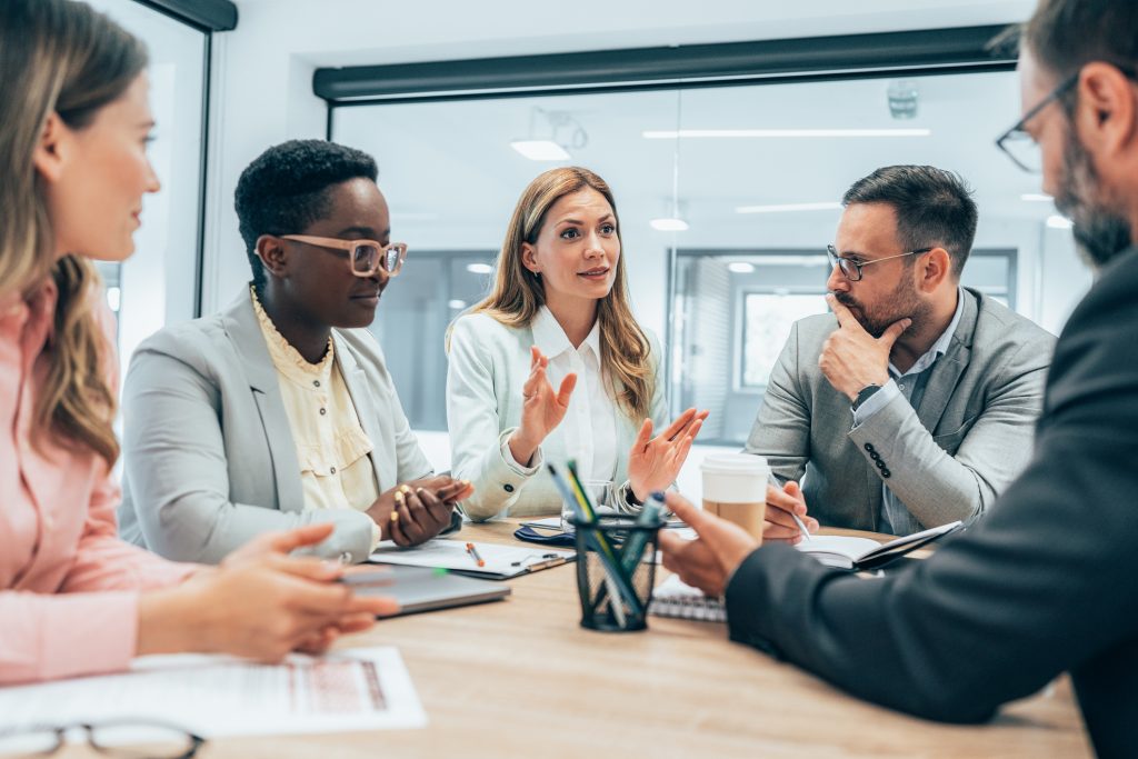 Cinq personnes en tenue de travail sont assises autour d'une table de conférence, engagées dans une discussion, avec des documents, des carnets de notes et une tasse à café sur la table.