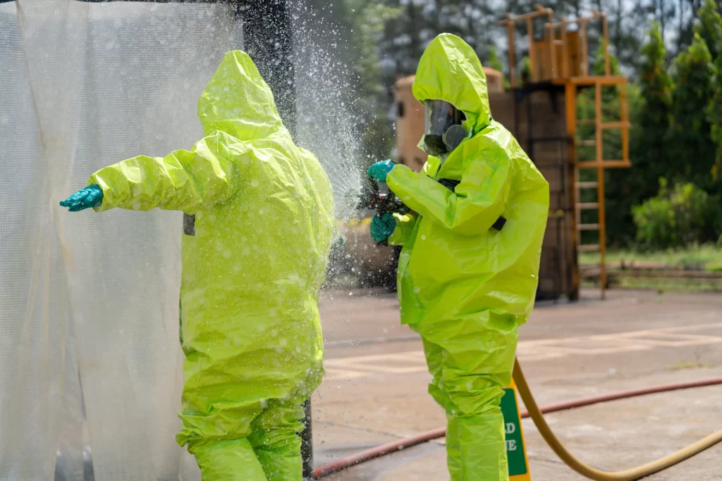 Deux personnes vêtues de combinaisons de protection jaune vif s'aspergent ou se décontaminent mutuellement avec un liquide à l'extérieur, près d'un équipement industriel.
