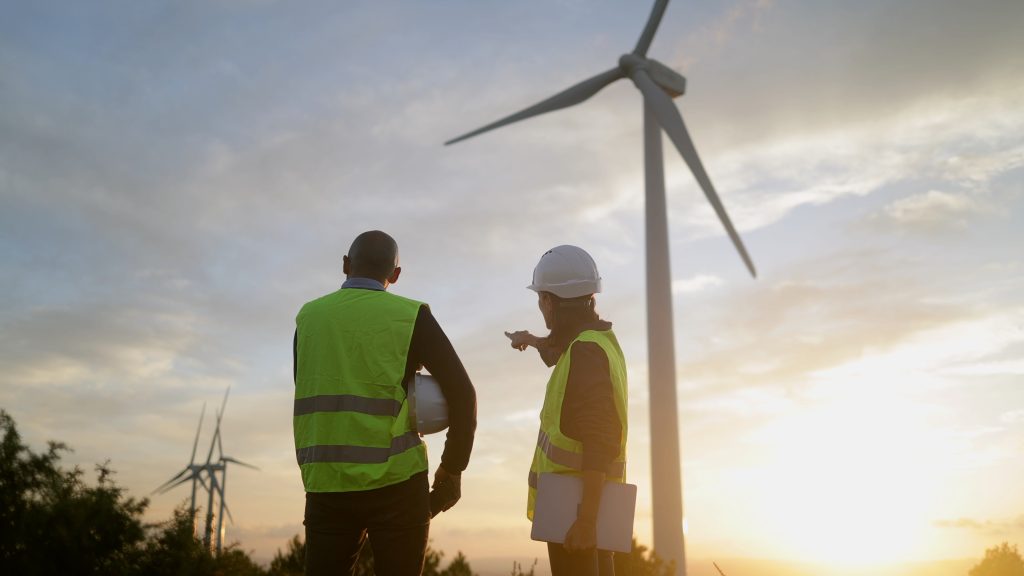 Deux ouvriers, vêtus de gilets de sécurité et coiffés de casques, se tiennent près d'éoliennes au coucher du soleil, en train de discuter ou d'inspecter le site.