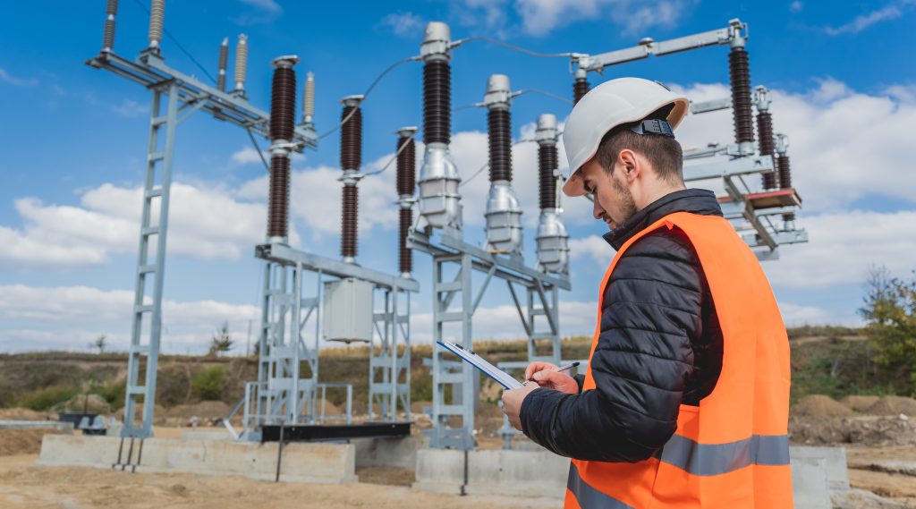 Un ingénieur portant un casque de chantier et un gilet de sécurité, en train de prendre des notes sur un bloc-notes devant un poste électrique.
