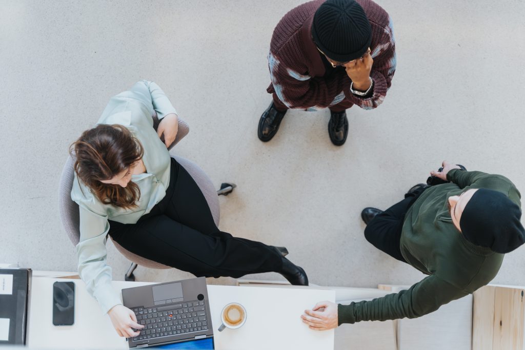 Trois personnes sont rassemblées autour d'une petite table ; l'une d'entre elles est assise devant un ordinateur portable, tandis que les deux autres se tiennent debout à proximité.